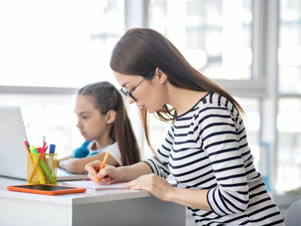 Block scheduling is one of the best productivity tips for moms, like this productive mom who is writing on a notepad while her daughter sits beside her, working on the computer.